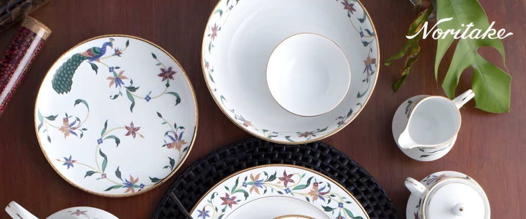 A table setting with Noritake Bountiful Garden collection dinnerware. The collection features a pattern of colorful peacocks and flowers. The image includes plates, bowls, and pitchers.